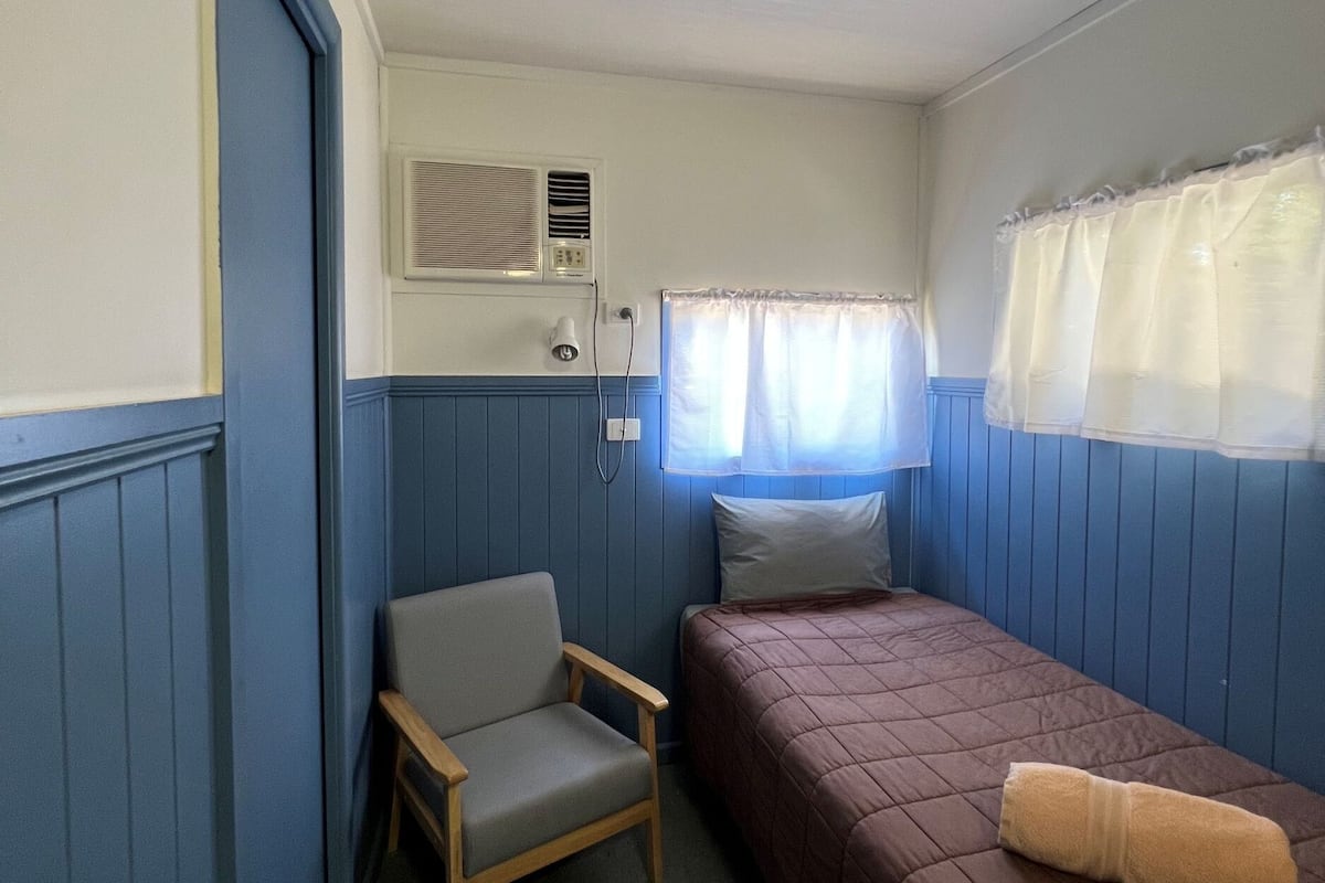 Eine kleine, einfache Unterkunft mit einem Bett, einem Sessel, einem Fenster mit Vorhängen und einer Wand-Klimaanlage.