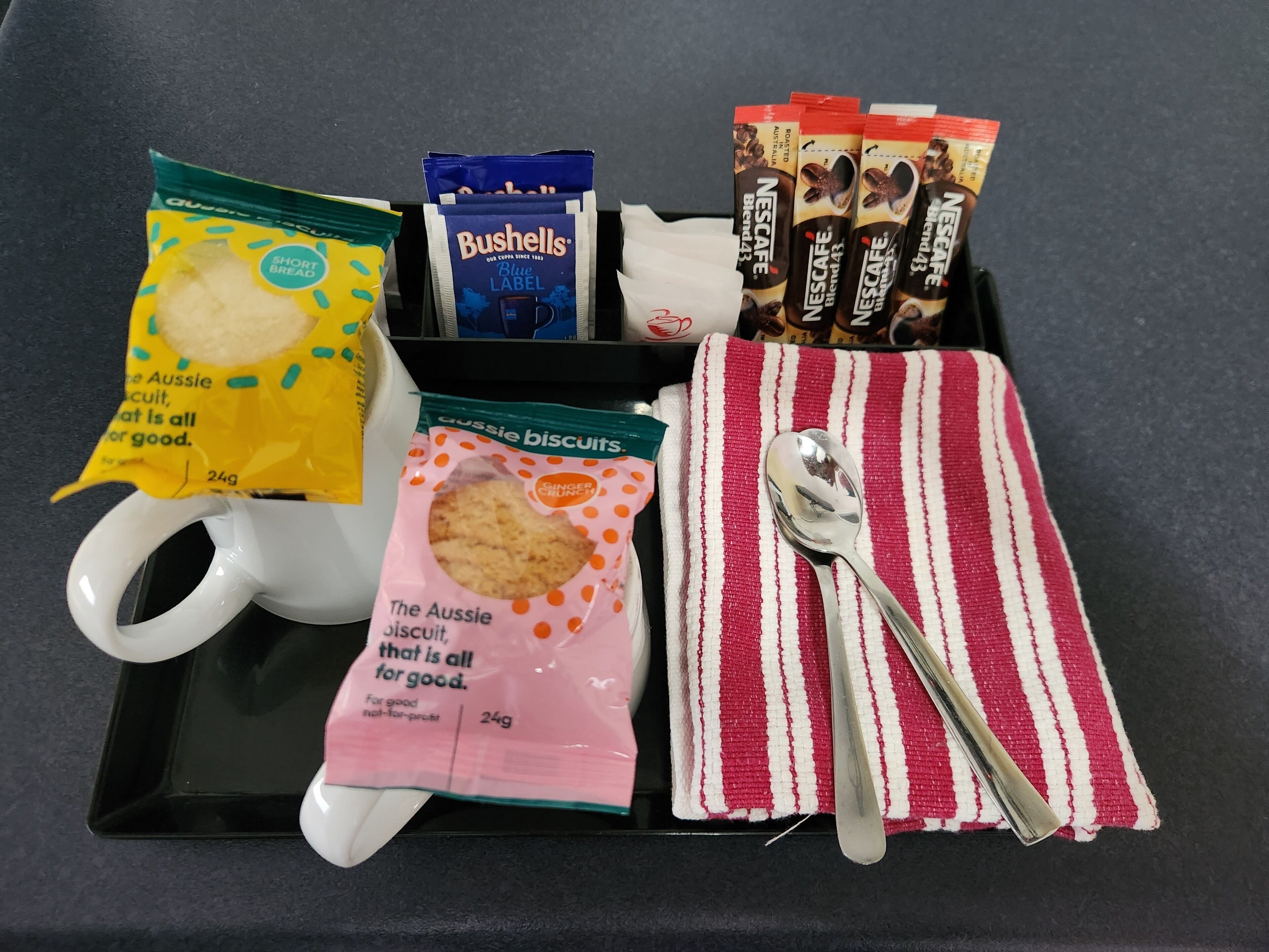 A tray with a white mug, a packet of shortbread biscuits, a packet of Bushells Blue Label coffee, a packet of The Aussie biscuit, and a striped cloth with a spoon on it.
