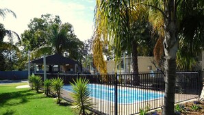 A swimming pool surrounded by a black fence, with a covered pavilion and palm trees in the background.