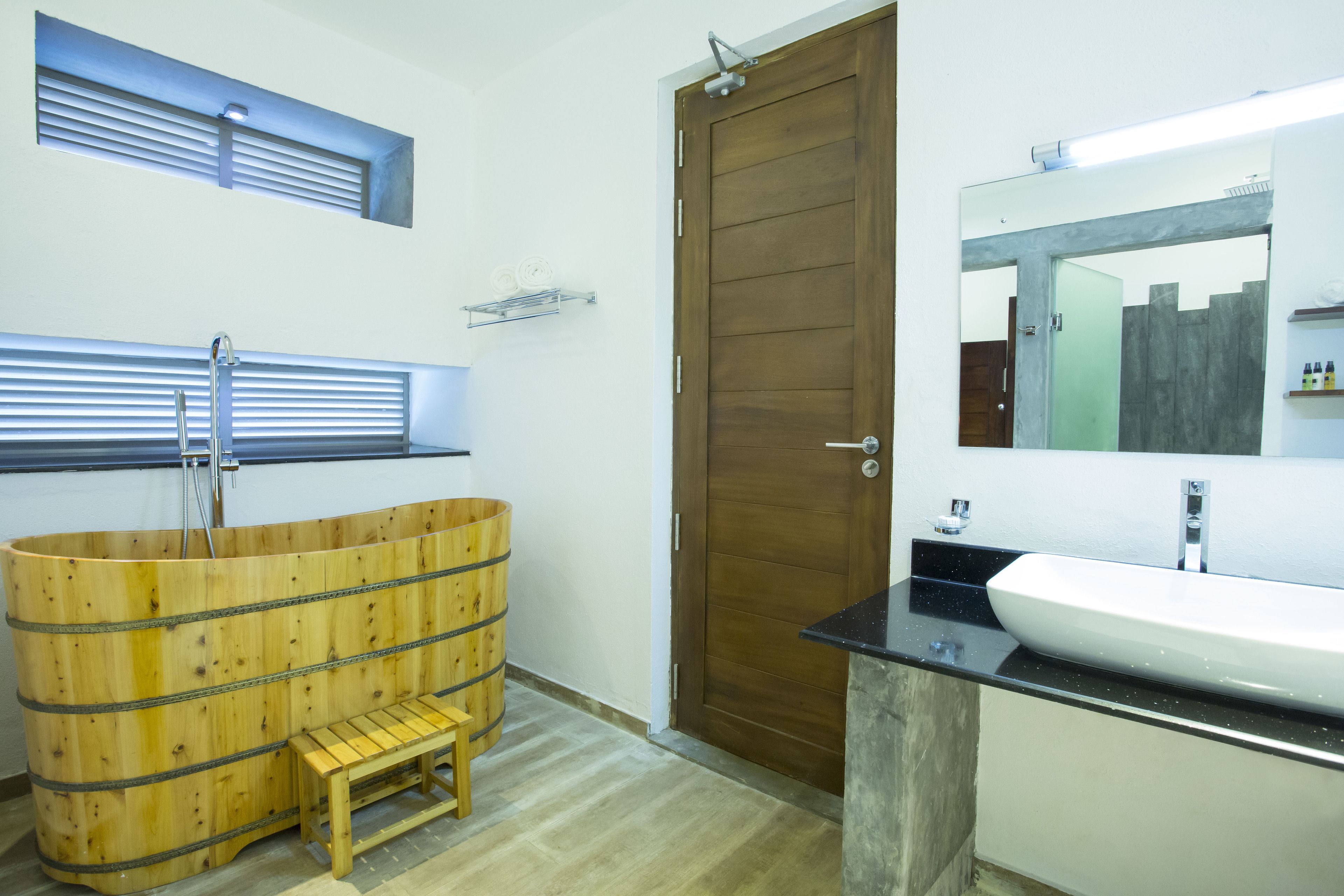 A bathroom with a wooden bathtub, a sink, a mirror, and a door.