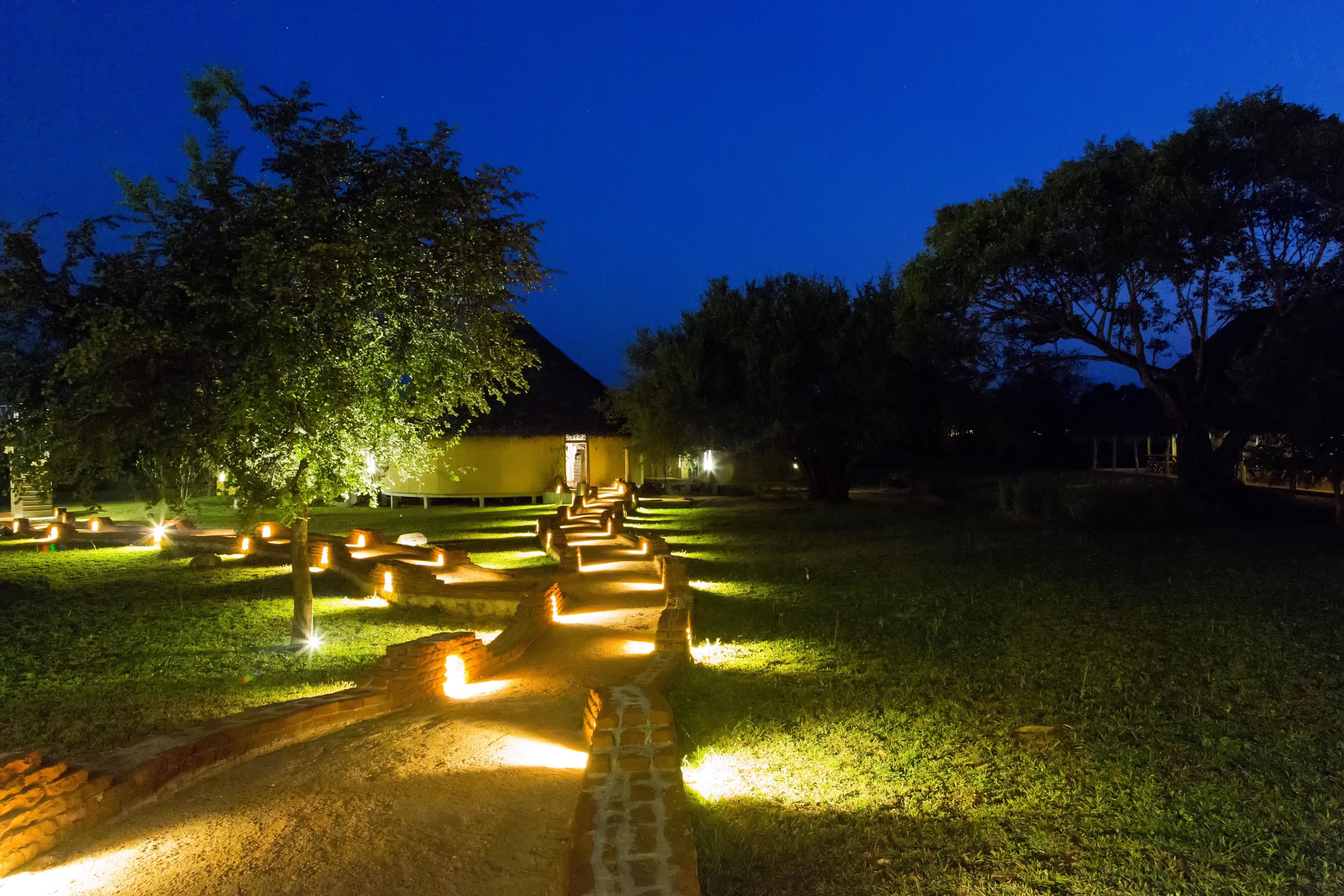 Un chemin bien éclairé mène à un bâtiment doté d’un toit en pente, entouré d’arbres et illuminé par des projecteurs au sol.
