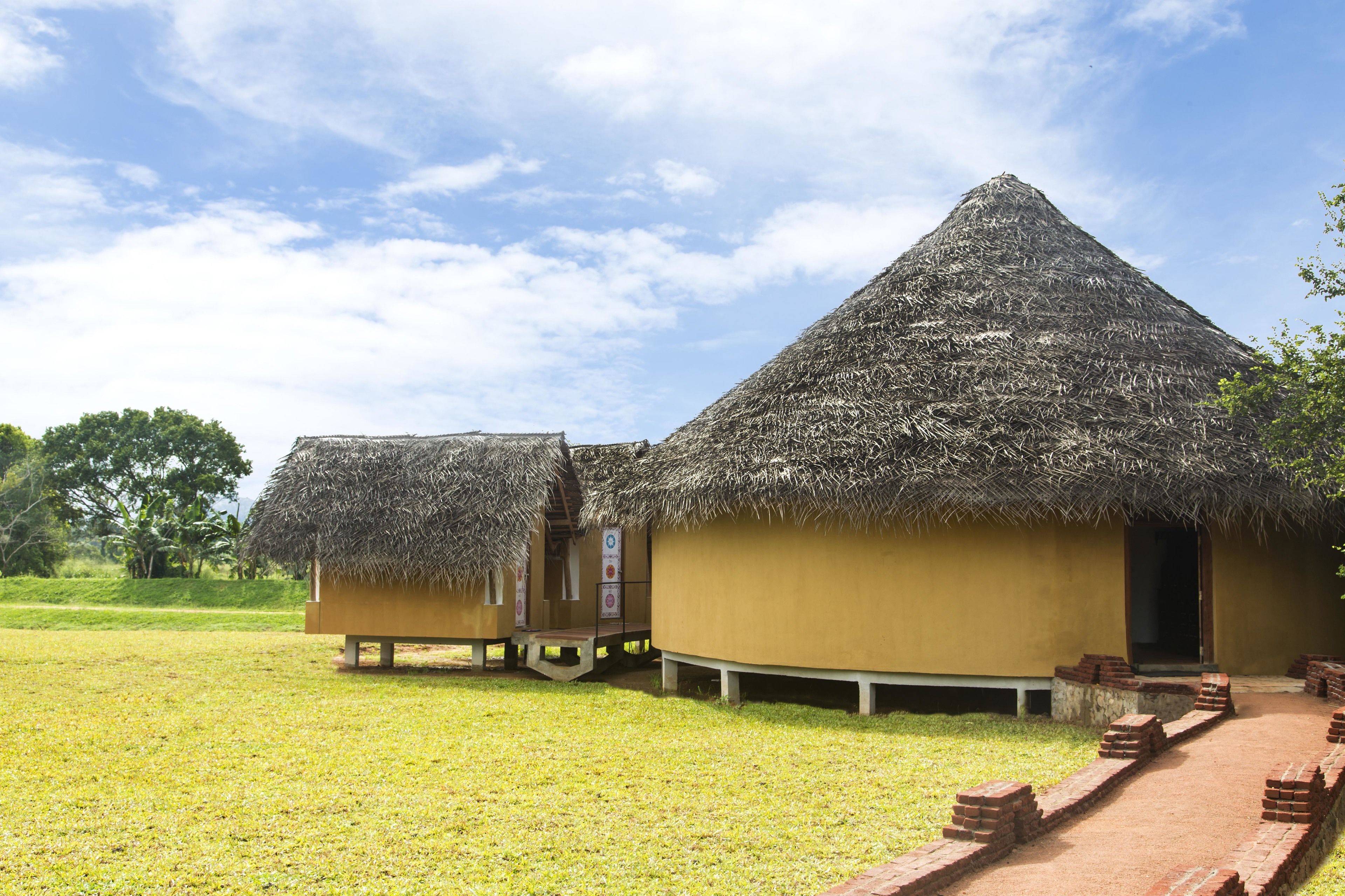 A traditional thatched-roof structure with a flat roof, surrounded by a grassy area and a brick pathway leading to the entrance.