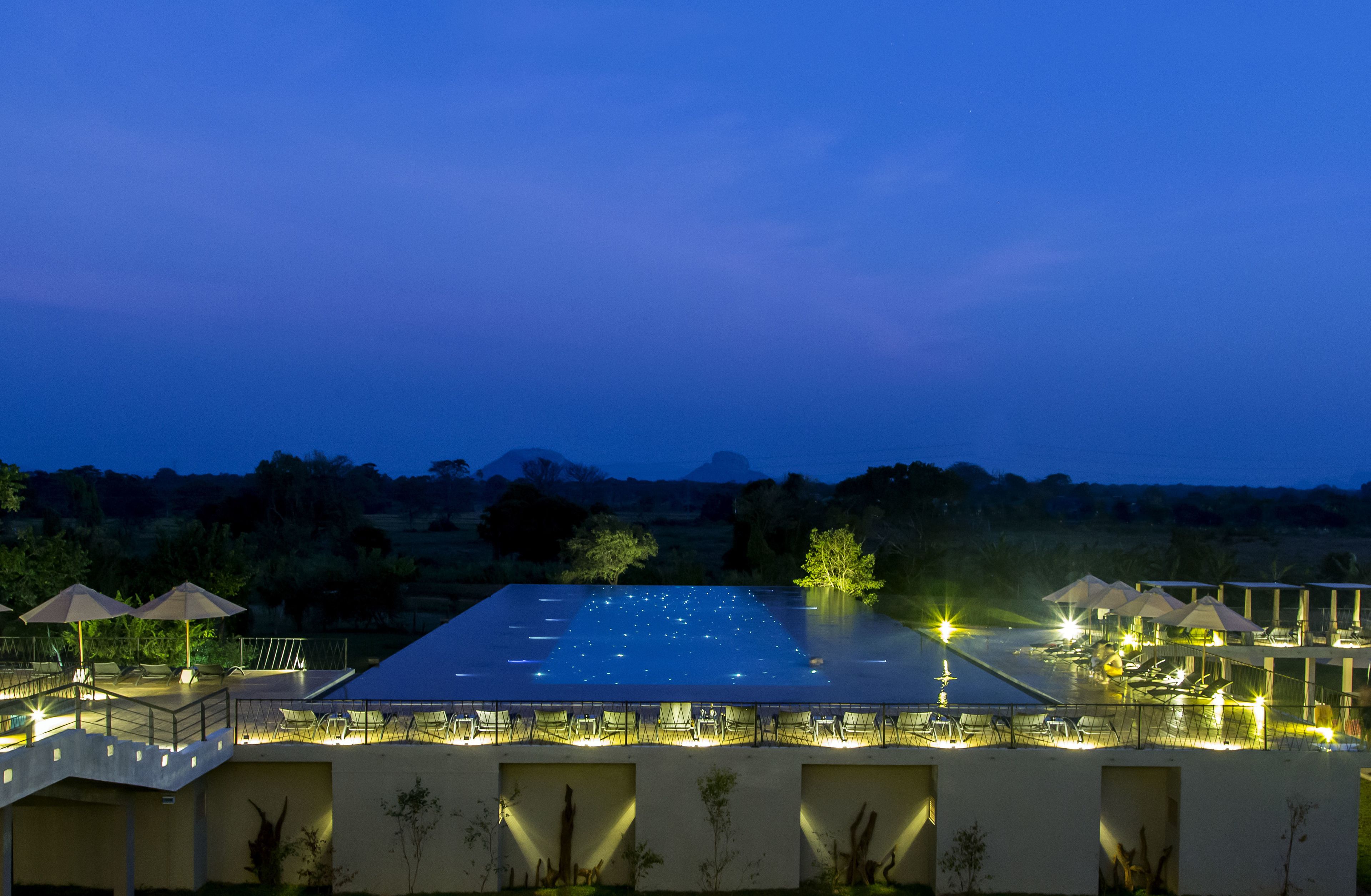 A large outdoor swimming pool at dusk with surrounding lounge areas and a view of distant hills.