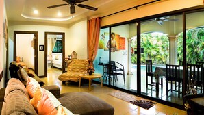 A spacious living area with a sofa, a coffee table, and a dining table with chairs. There is a sliding glass door leading to an outdoor patio with a view of greenery. - Coconut Paradise Villas (Rawai)