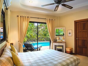 A bedroom with a bed, a desk, a chair, a window with curtains, a ceiling fan, and a view of a swimming pool.