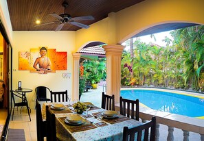 A dining area with a table set for four, a Buddha painting on the wall, and a view of a swimming pool with tropical plants.