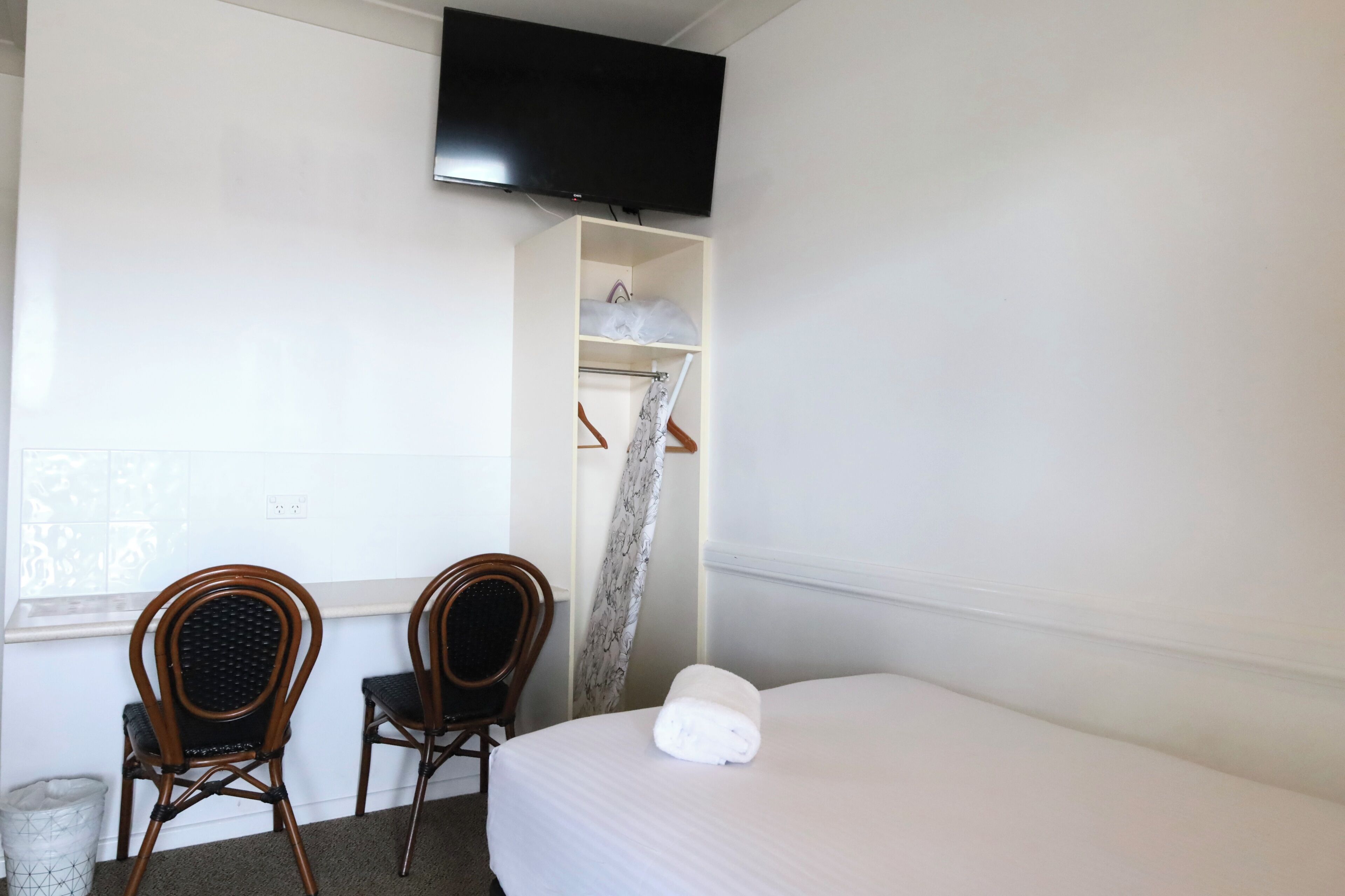 A hotel room with a bed, two wooden chairs, a TV mounted on the wall, a window with patterned curtains, and a small shelf with folded clothes.