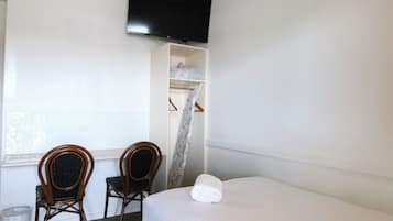 A hotel room with a bed, two wooden chairs, a TV mounted on the wall, a window with patterned curtains, and a small shelf with folded clothes.