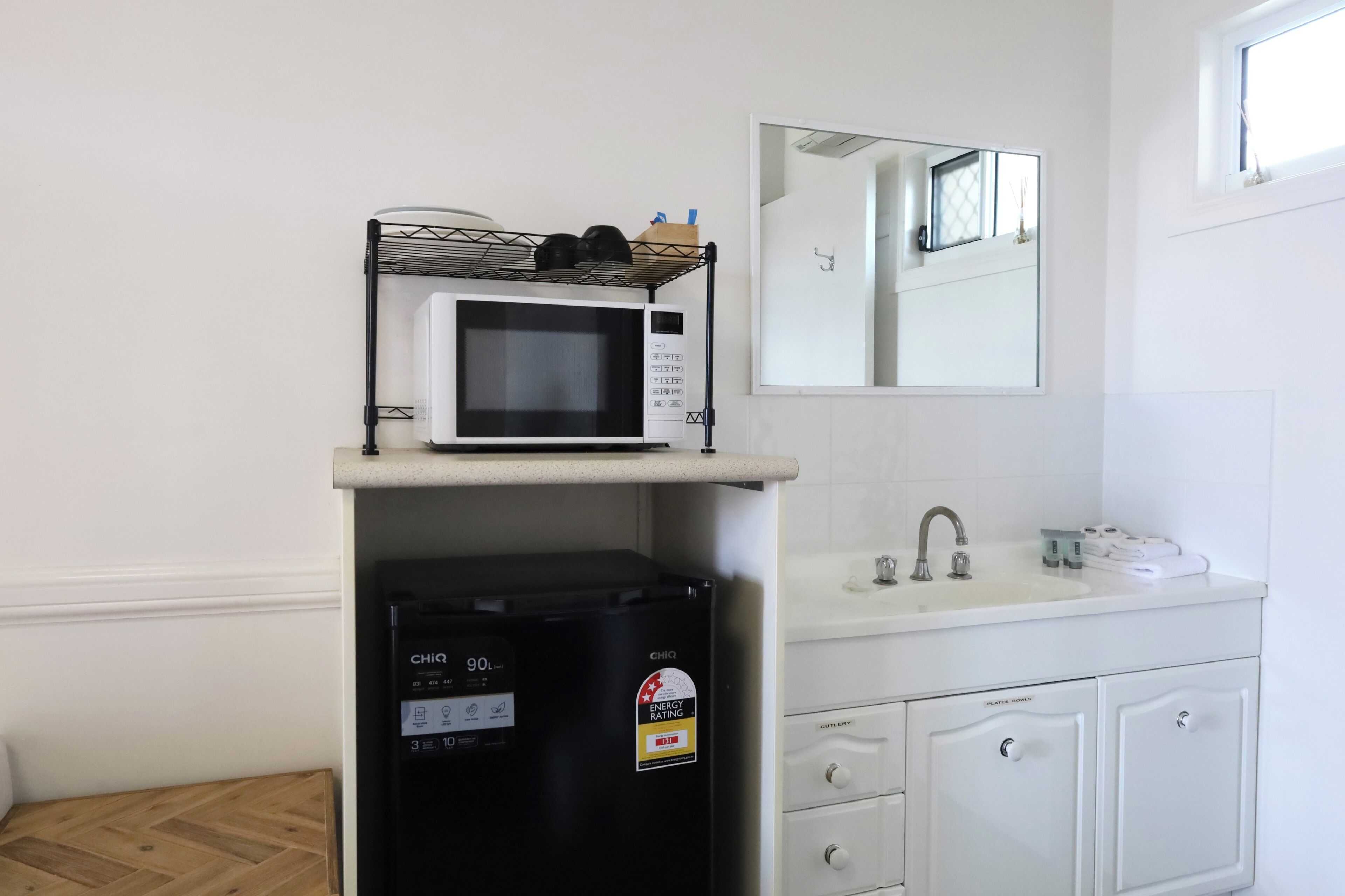 Een keukentje met een magnetron op een plank, een zwarte koelkast eronder, een spoelbak met een spiegel erboven en een raam voor natuurlijk licht.