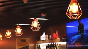 Een barruimte met hangende geometrische lampen, een televisiescherm en een plank met verschillende flessen.