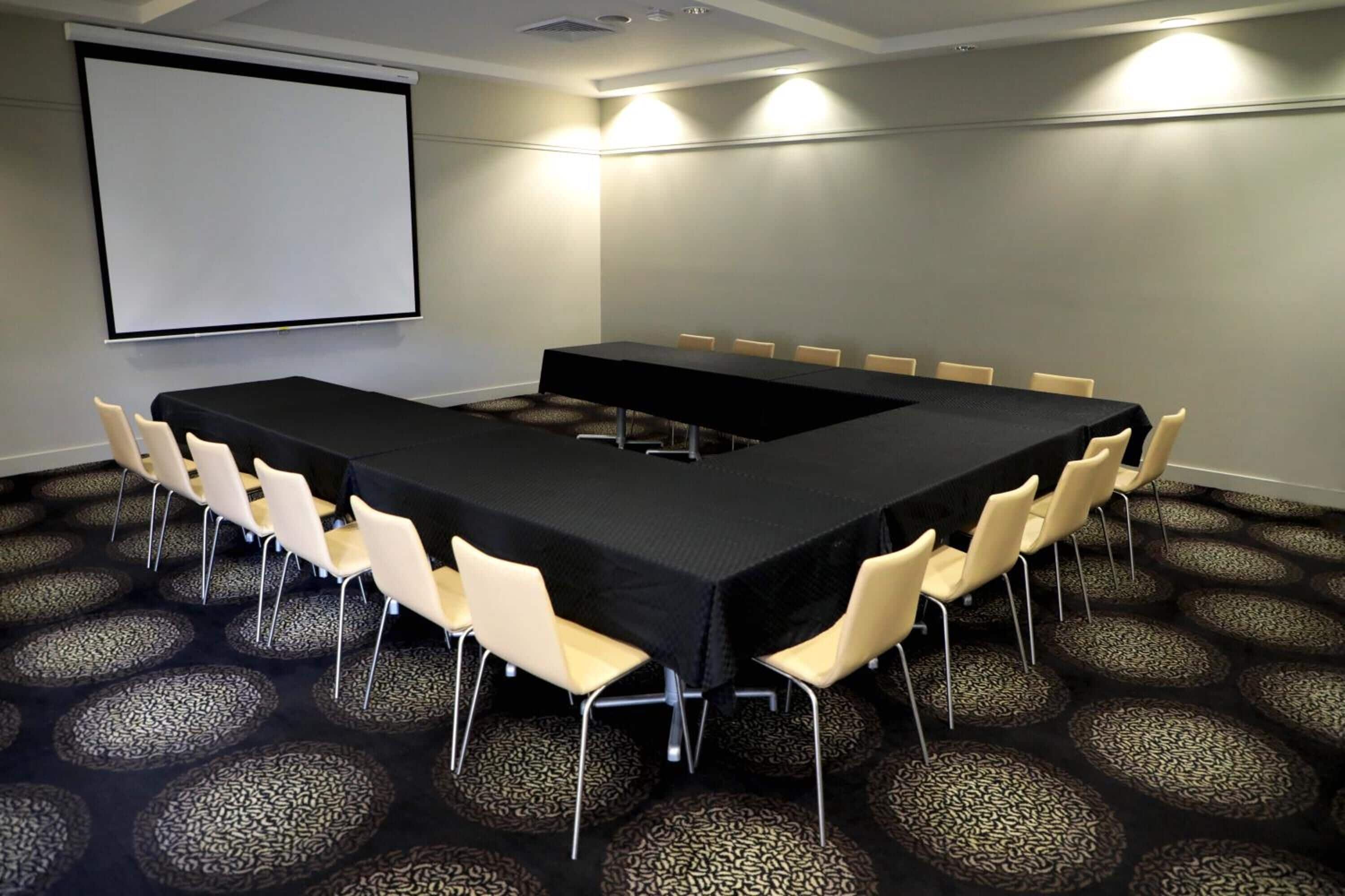 A conference room with a long table, chairs, a projector screen, and patterned carpet.