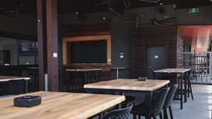 Een restaurantinterieur met meerdere houten tafels en zwarte stoelen, een groot raam met zonwering en een plafondventilator.