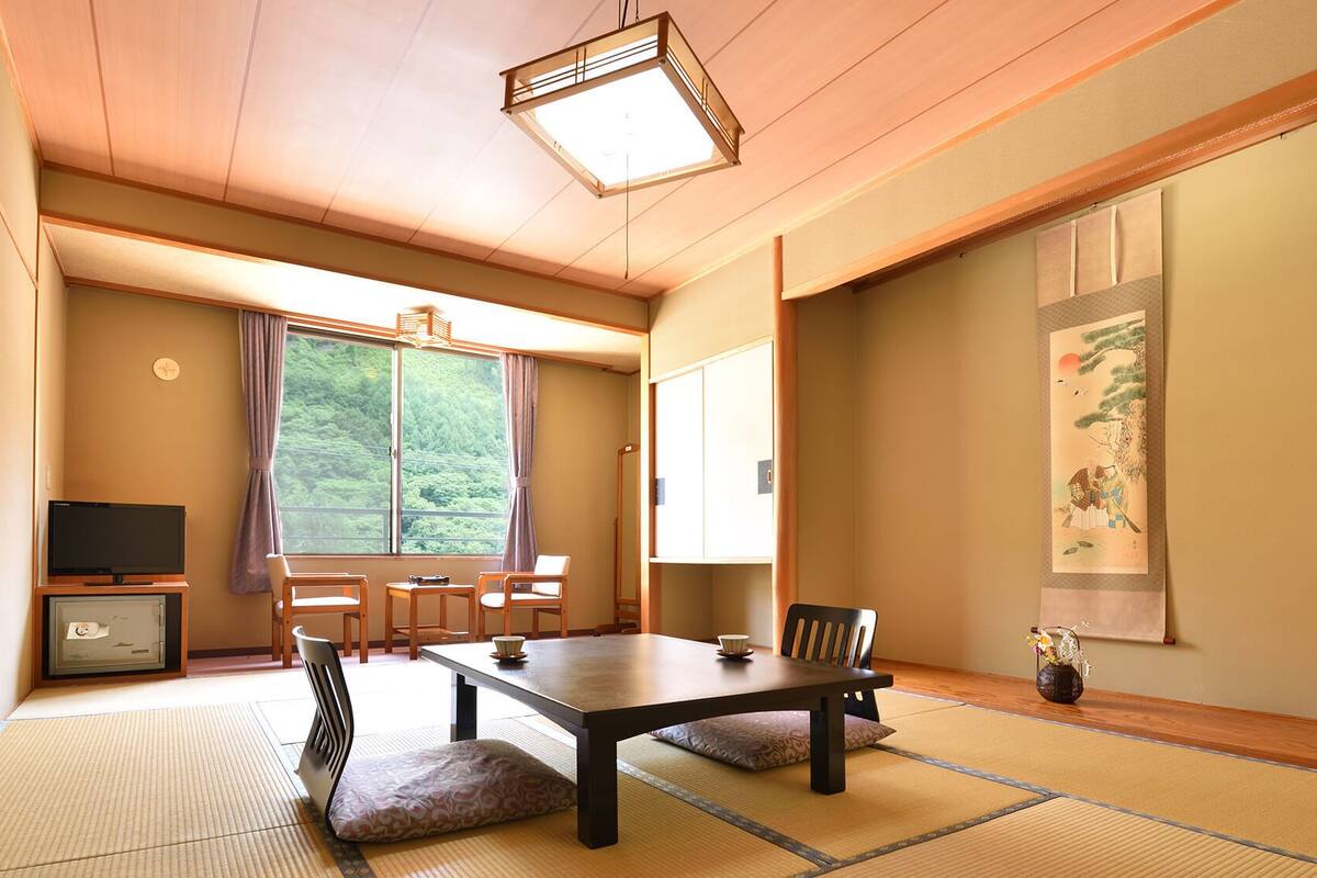A traditional Japanese room with tatami flooring, a low table, chairs, a television, and a view of a forest through a large window.