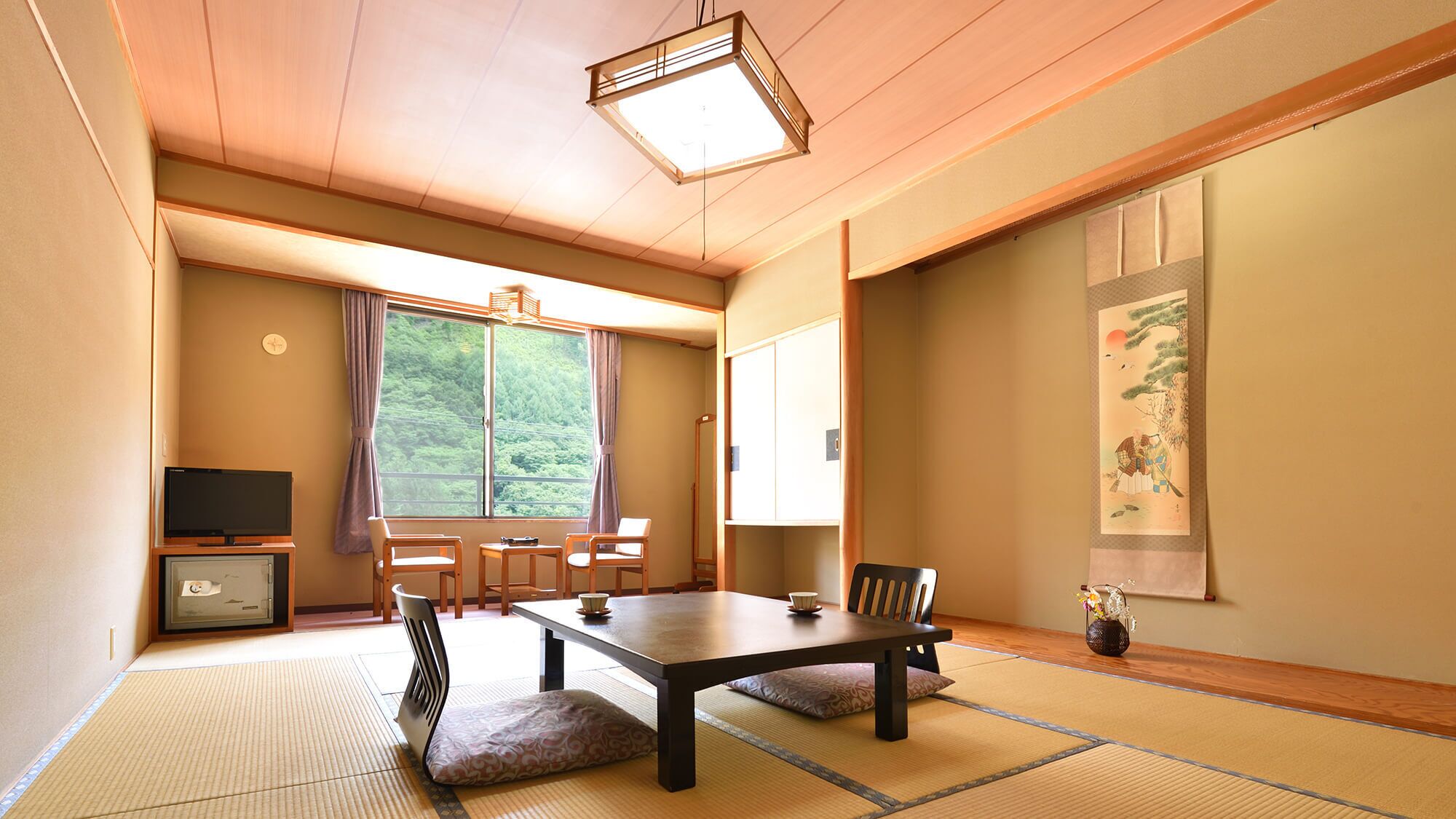 A traditional Japanese room with tatami flooring, a low table, chairs, a television, and a large window with a view of greenery.
