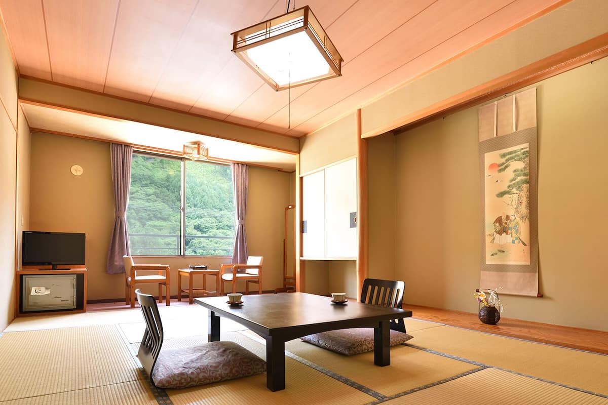 A traditional Japanese room with tatami flooring, a low table, chairs, a television, and a large window with a view of greenery.
