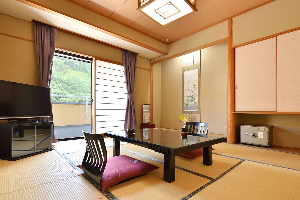 A traditional Japanese room with tatami flooring, a television, a table with chairs, and a sliding door leading to a balcony.