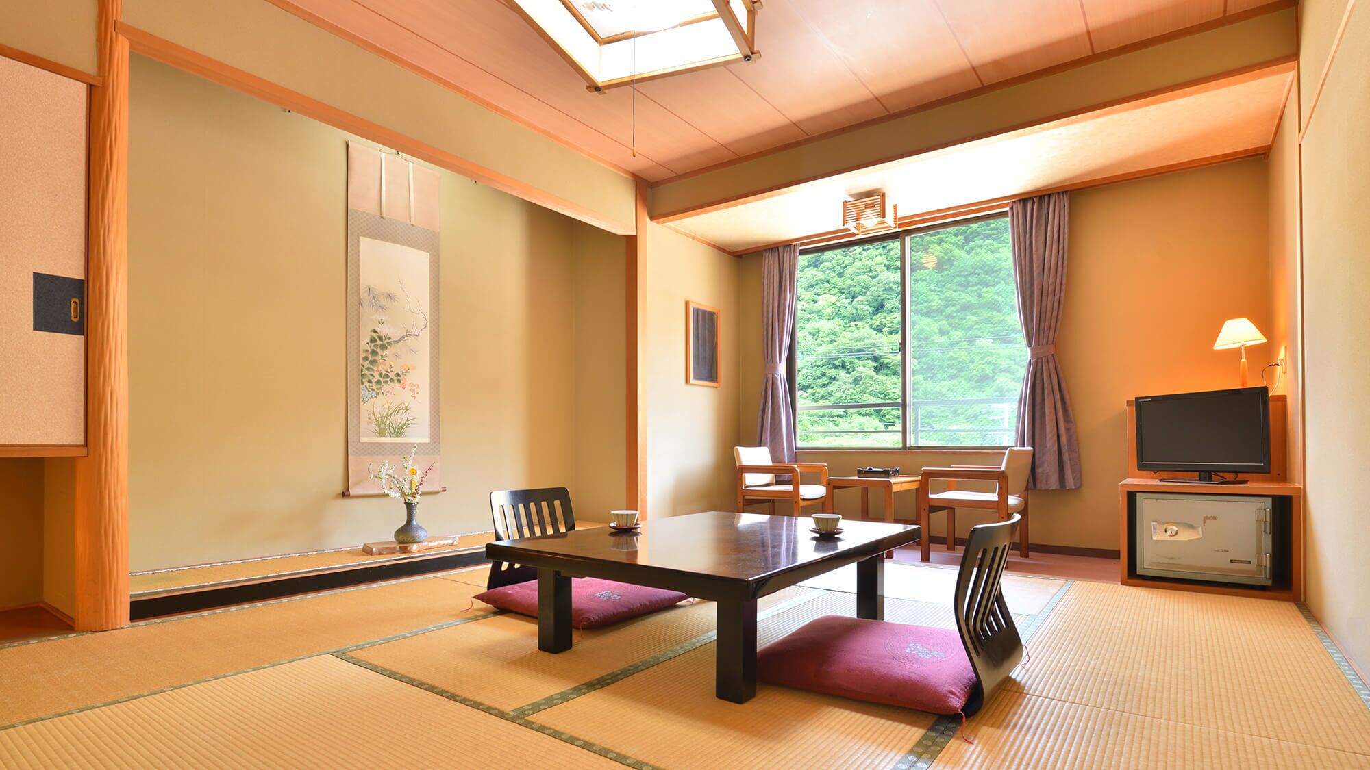 A traditional Japanese room with tatami flooring, a low table, chairs, a TV, and a window with a view of greenery.