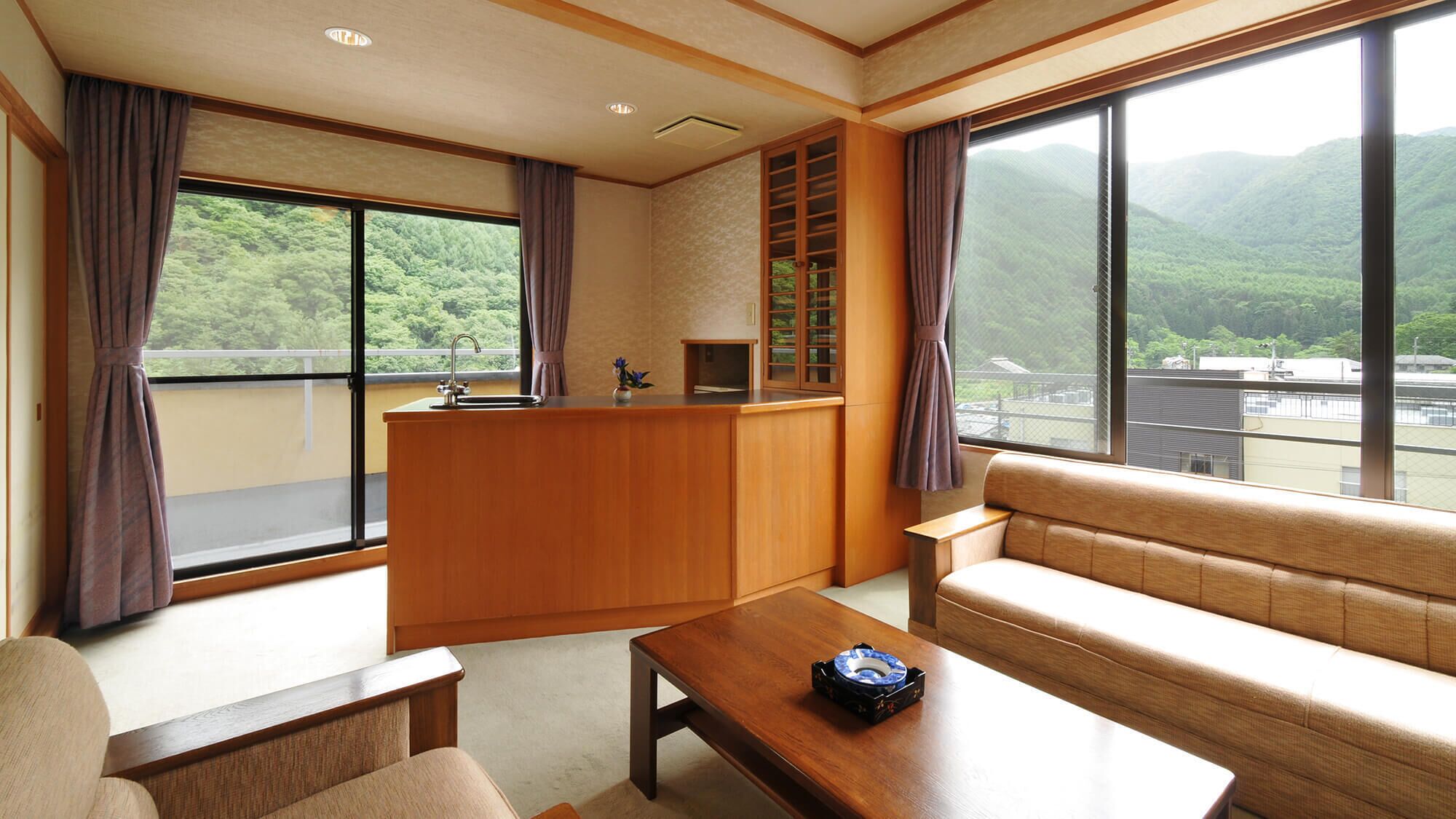 A room with a sofa, a small table, a wooden counter with a sink, and large windows offering a view of green hills.