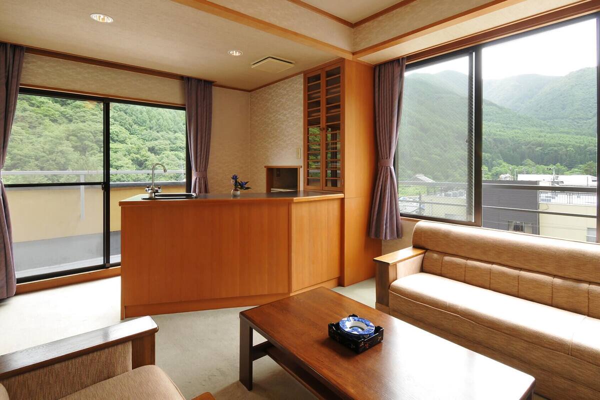 A room with a sofa, a small table, a wooden counter with a sink, and large windows offering a view of green hills.