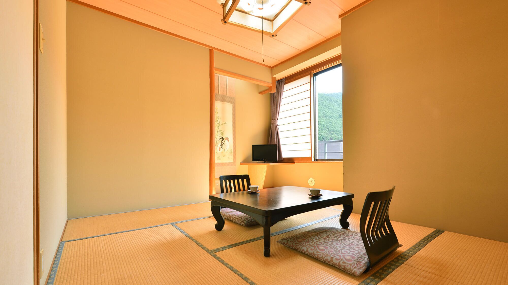 A tatami mat room with a low table and chairs, a television, and a window with blinds.