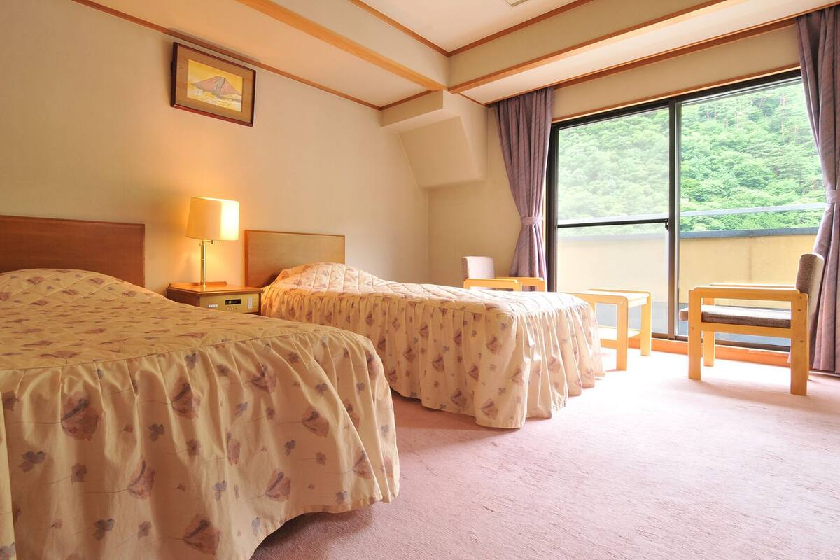 A hotel room with two beds, a nightstand, a lamp, a chair, and a large window with a view of greenery.