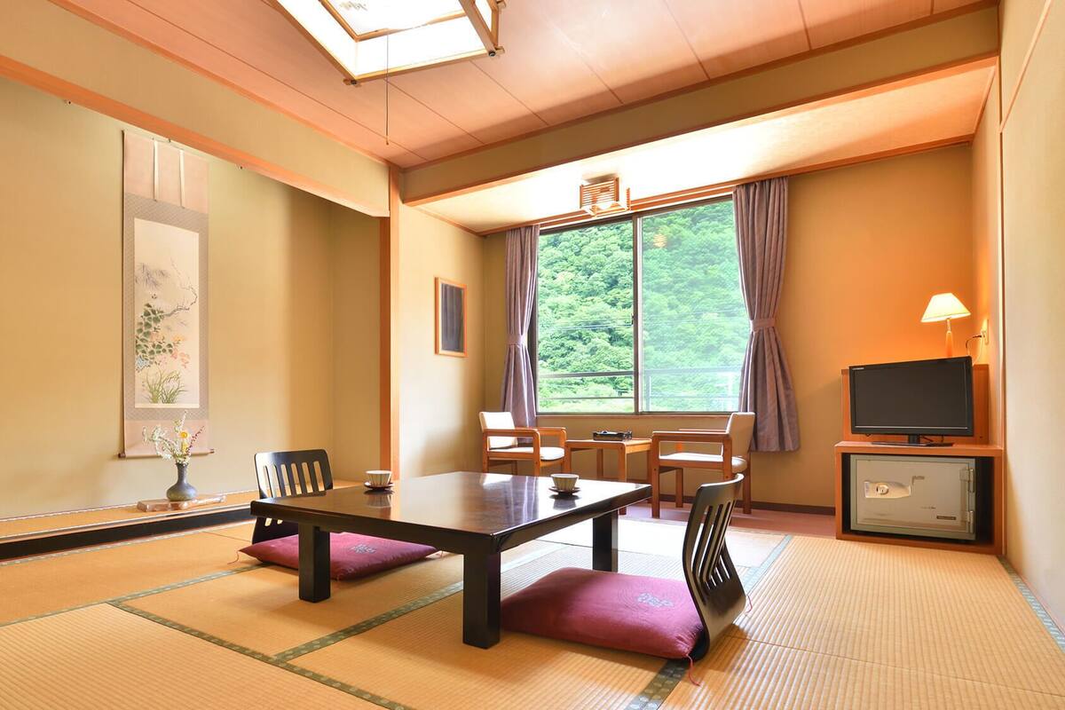 A traditional Japanese-style room with tatami flooring, a low table, chairs, a television, and a window with a view of greenery.