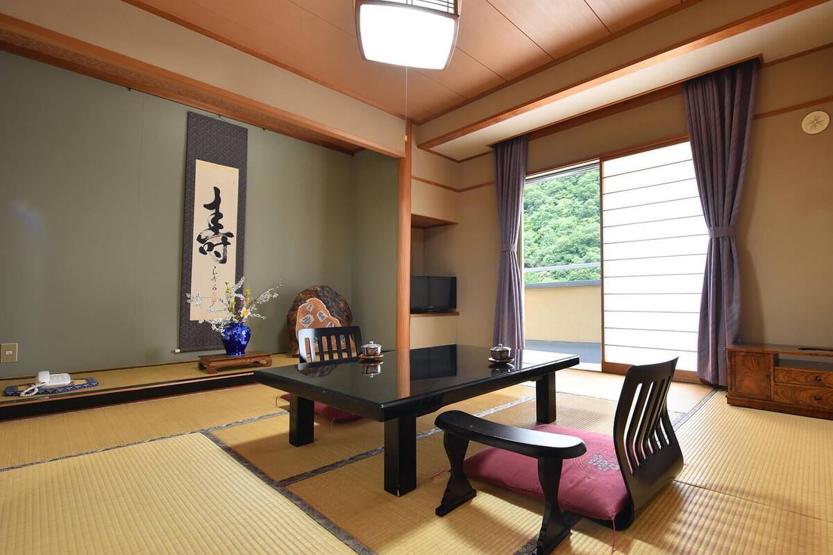 A traditional Japanese room with tatami flooring, a low table, a bench, a TV, and a sliding door with a view of greenery.