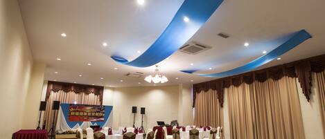 Un salón de banquetes con mesas redondas, sillas y un escenario con un cartel.