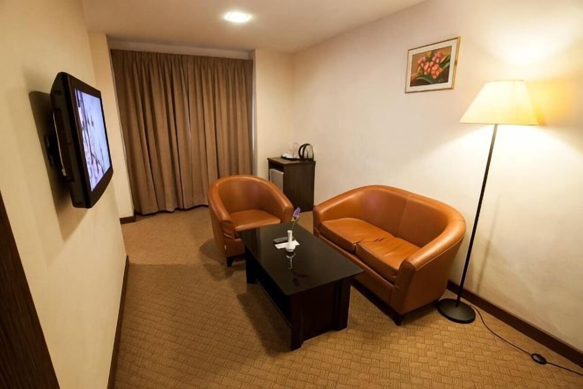 Habitación de hotel con sofá de cuero marrón, una mesita, televisor, lámpara y ventana con cortinas.