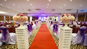 A banquet hall with a red carpet aisle, white chairs with purple sashes, and floral centerpieces.