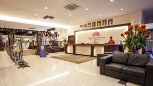 A hotel reception area with a reception desk, a receptionist, a sofa, a potted plant, and a clock on the wall.