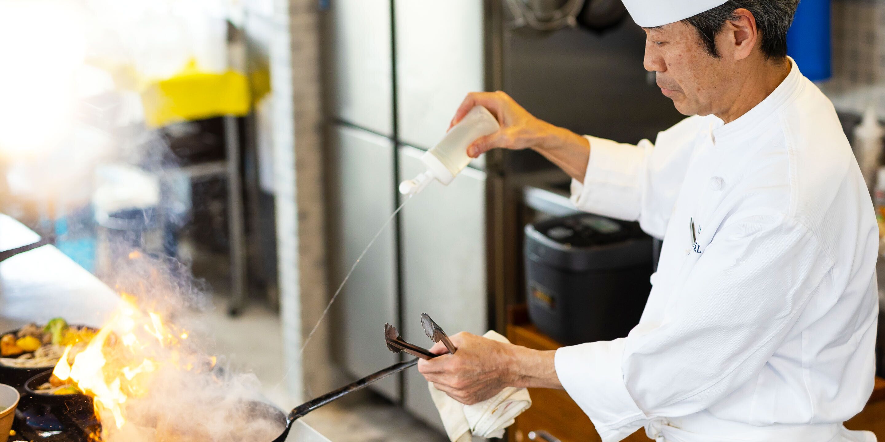 A chef cooking with a pan on a stove, using tongs and a bottle.