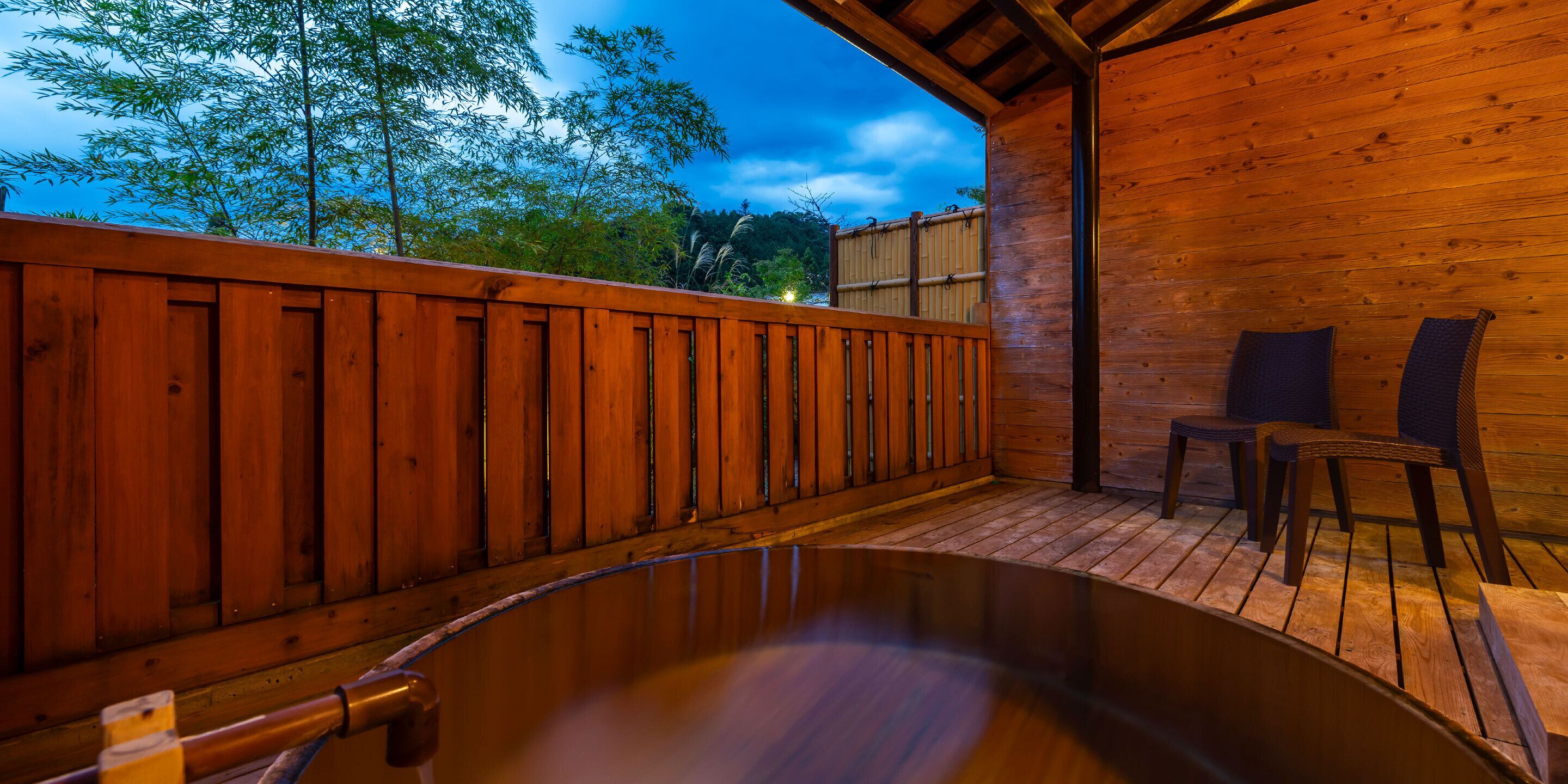 Una terraza de madera con un jacuzzi, sillas de madera y vistas a árboles y al cielo.
