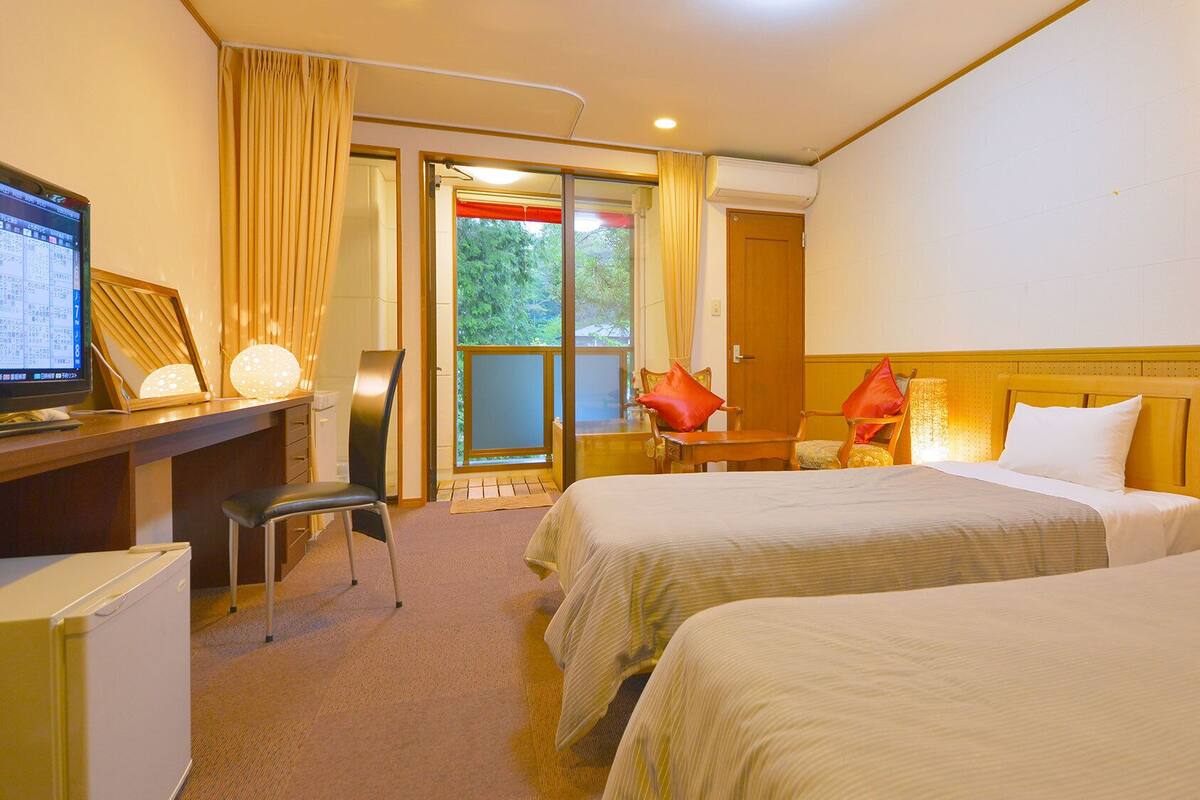 A hotel room with two beds, a desk with a computer, a chair, and a view of greenery outside.