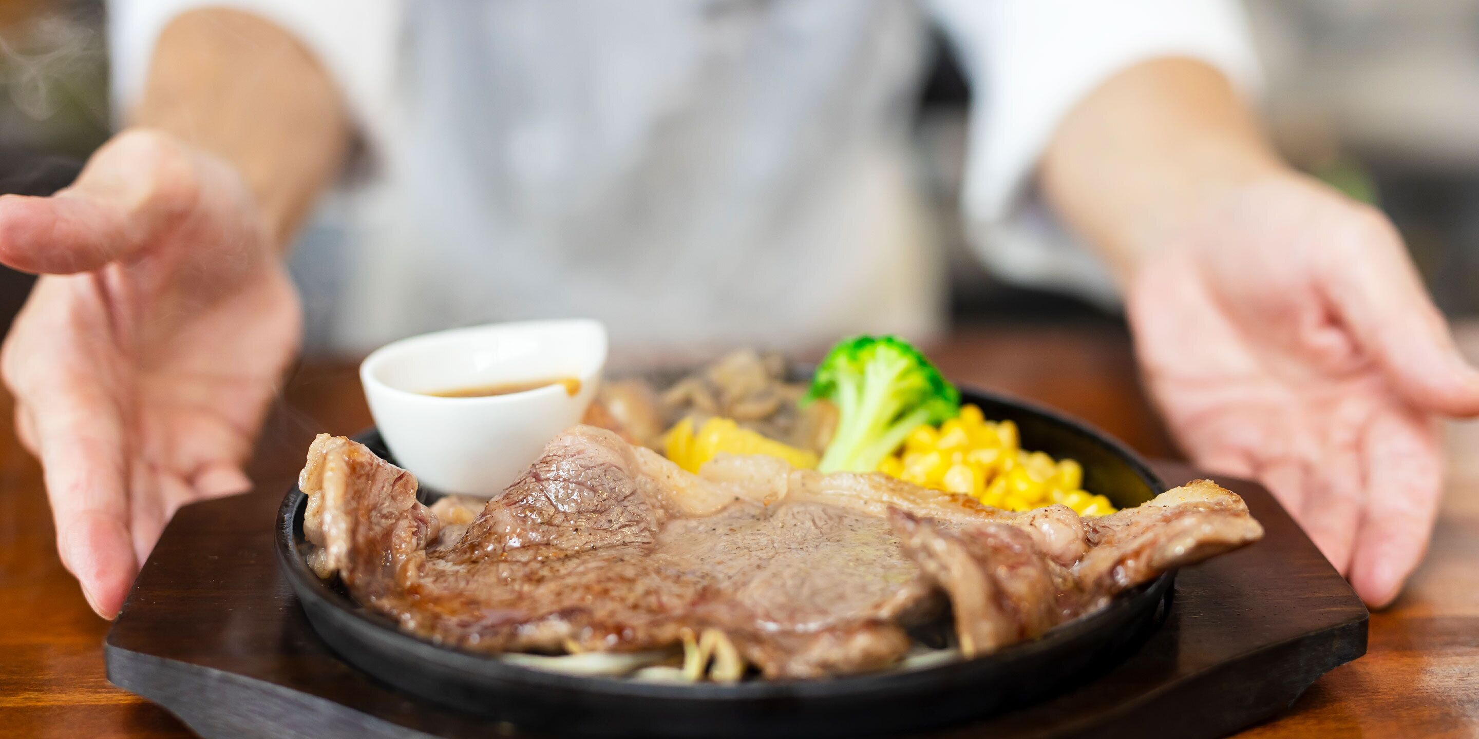 A plate of food with a piece of meat, corn, and broccoli, accompanied by a small bowl of sauce.