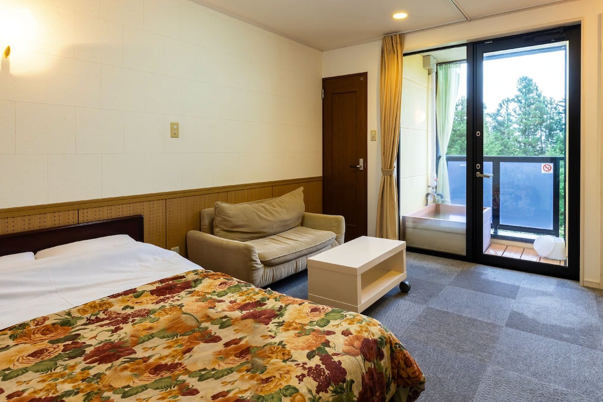A hotel room with a bed, a sofa, a small table, and a sliding glass door leading to a balcony.