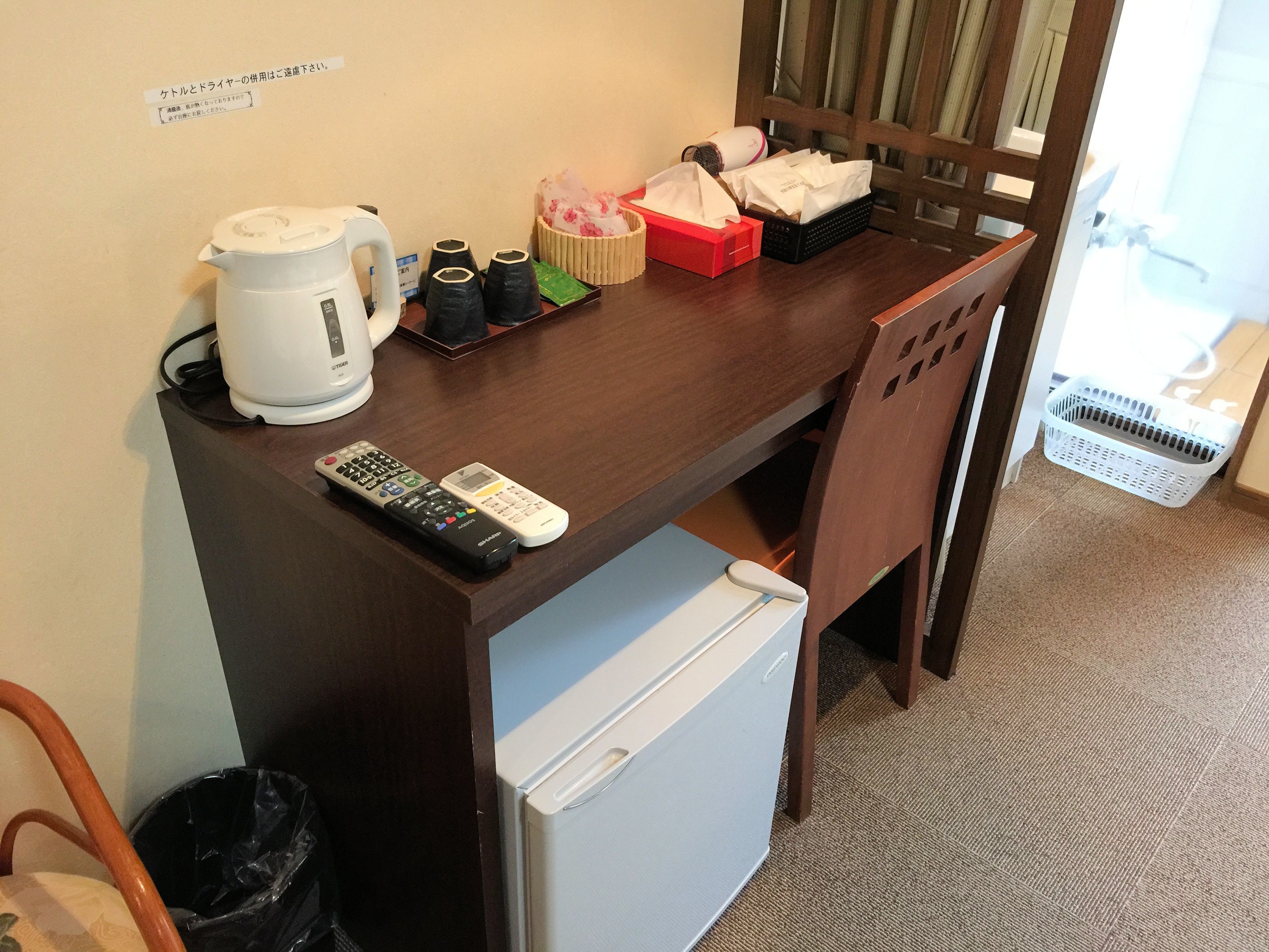 Habitación de hotel compacta con kitchenette, refrigerador, cafetera, microondas, hervidor de agua, basurero y una silla de madera.