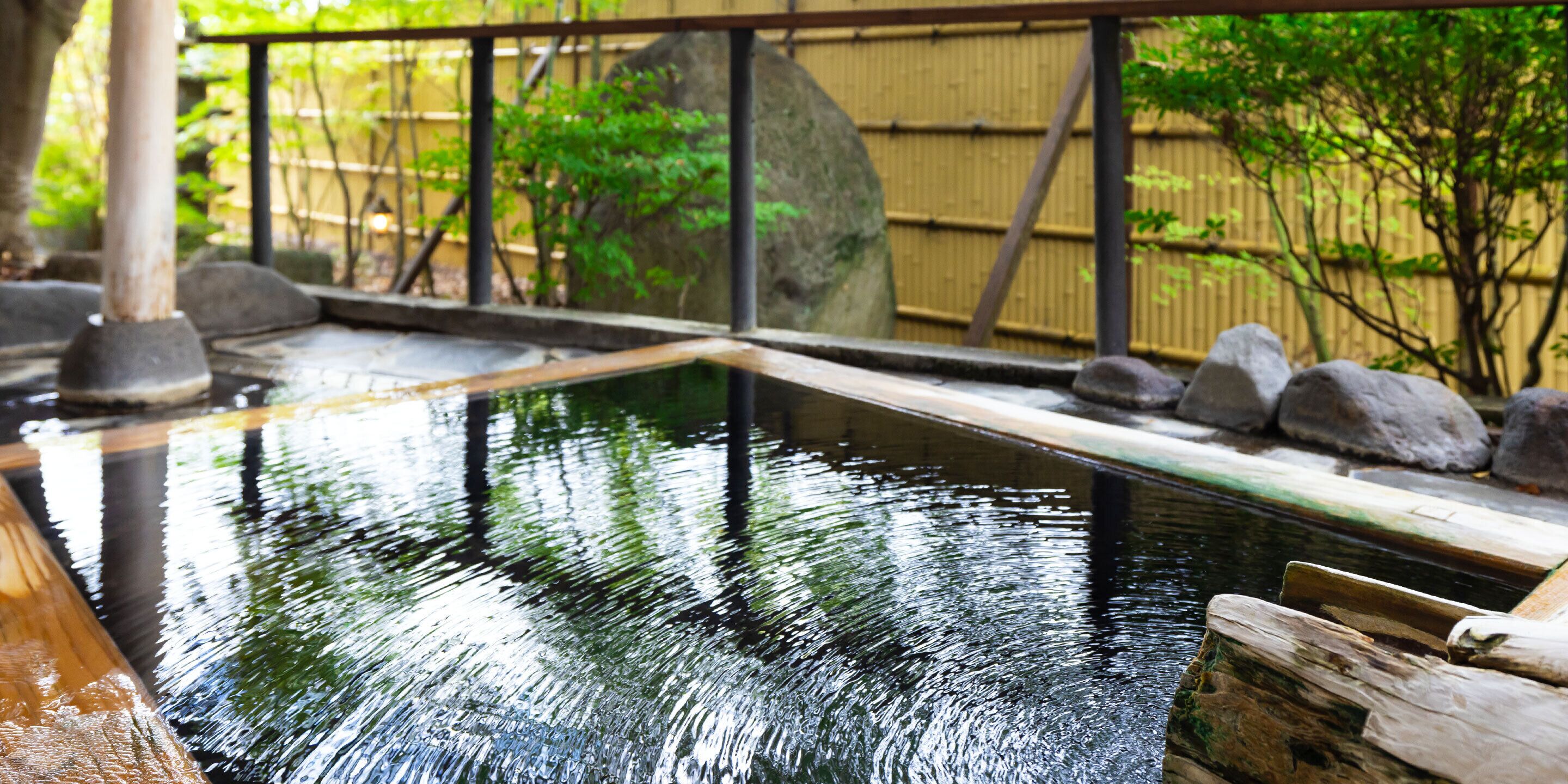 A traditional Japanese onsen with a wooden deck, a stone pathway, and a bamboo fence in the background.