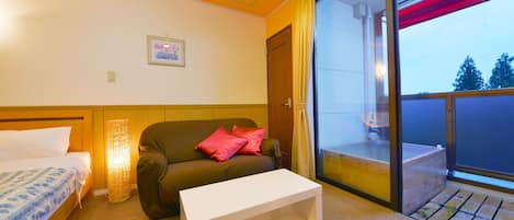 A hotel room with a bed, a sofa, a small table, a window with a view of trees, and a door.