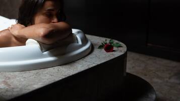 Una persona recostada con la cabeza en una bañera blanca en una habitación tenuemente iluminada, con una encimera de mármol y una pequeña rosa roja encima.