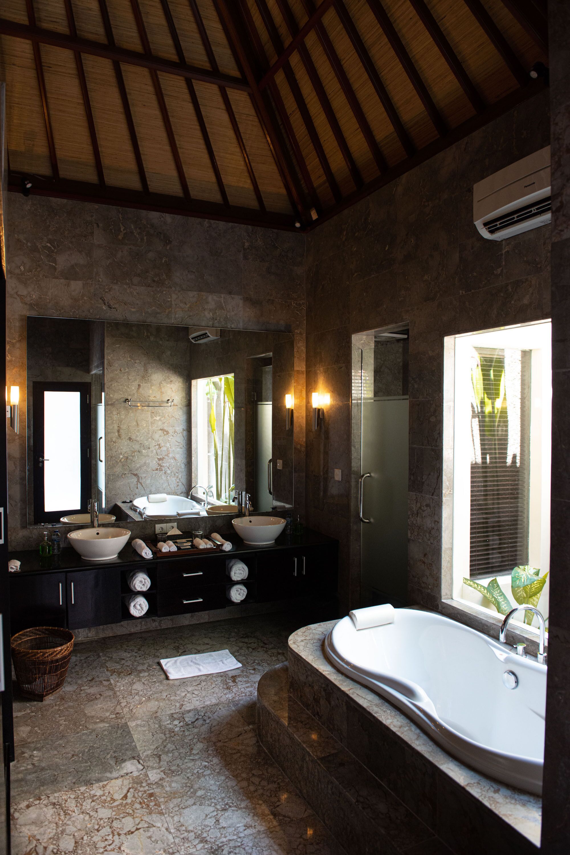 A bathroom with a large bathtub, two sinks, a mirror, and a window with a view of greenery.