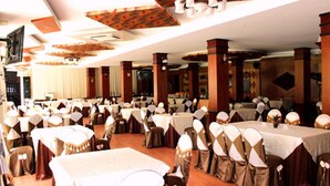 A banquet hall with multiple round tables covered with white tablecloths, each surrounded by chairs with white and brown covers. The hall features wooden columns and a patterned ceiling design. - Cuu Long Hotel (Can Tho)