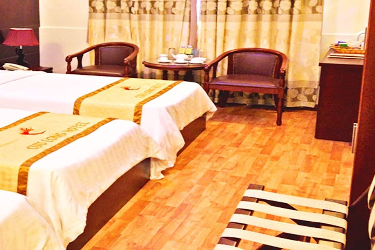 Habitación de hotel con tres camas, cada una con la etiqueta 'Deluxe' y el logotipo de 'Long Hotel'. El suelo es de madera, hay una mesita con una lámpara y dos sillas.