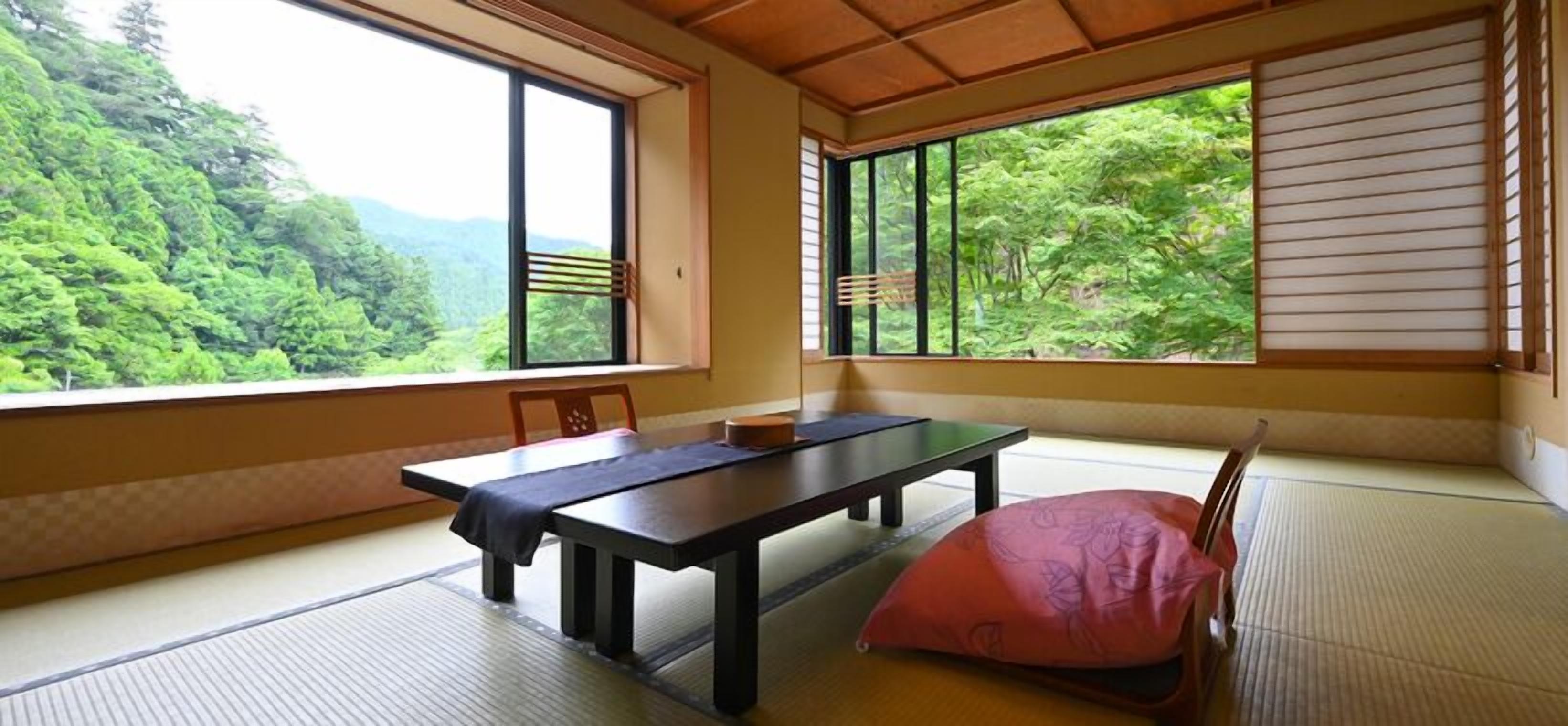 Ein Raum mit Tatami-Matten, einem Tisch und Stühlen, großen Fenstern mit Holzrahmen und Blick auf ein bewaldetes Gebiet.