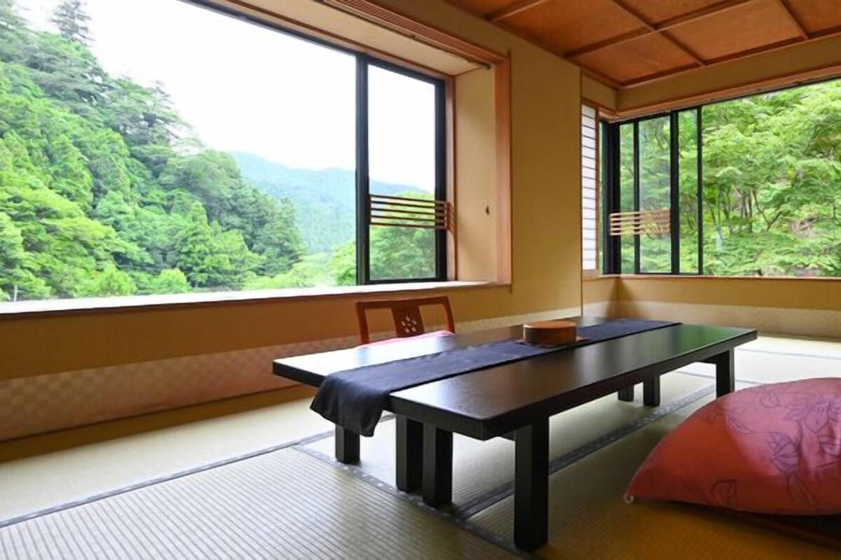 A tatami mat room with a table and chairs, large windows with wooden frames, and a view of a forested area.