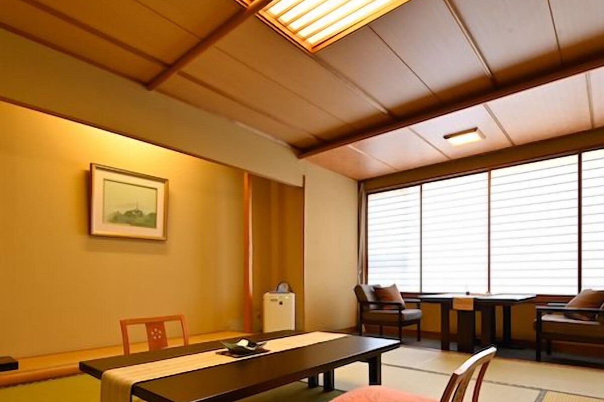 Een kamer met tatami matten, een lage tafel, stoelen, een klein kastje en een raam met jaloezieën.