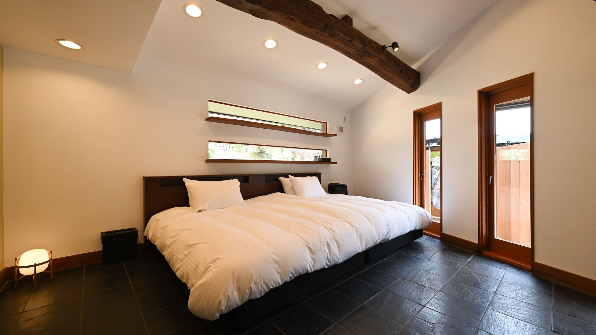 Une chambre avec un grand lit, des poutres en bois, des spots encastrés au plafond et une vue sur l’extérieur, visible à travers une porte.