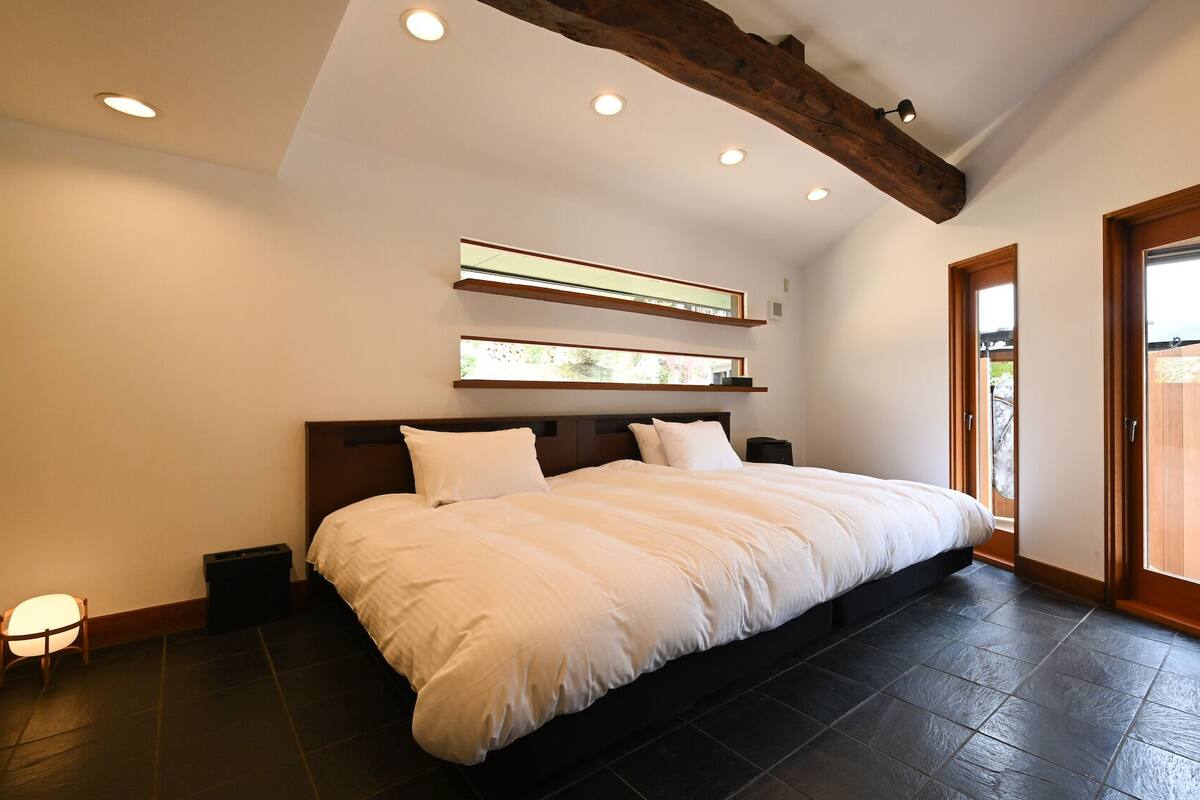 A bedroom with a large bed, wooden beams, recessed ceiling lights, and a view through a door.