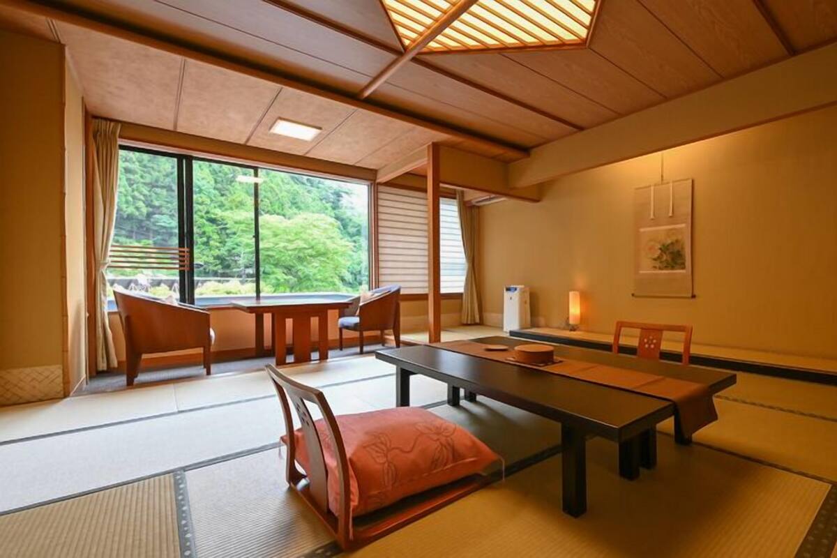 A traditional Japanese room with tatami flooring, a low table, chairs, a sliding door, and a view of greenery outside.