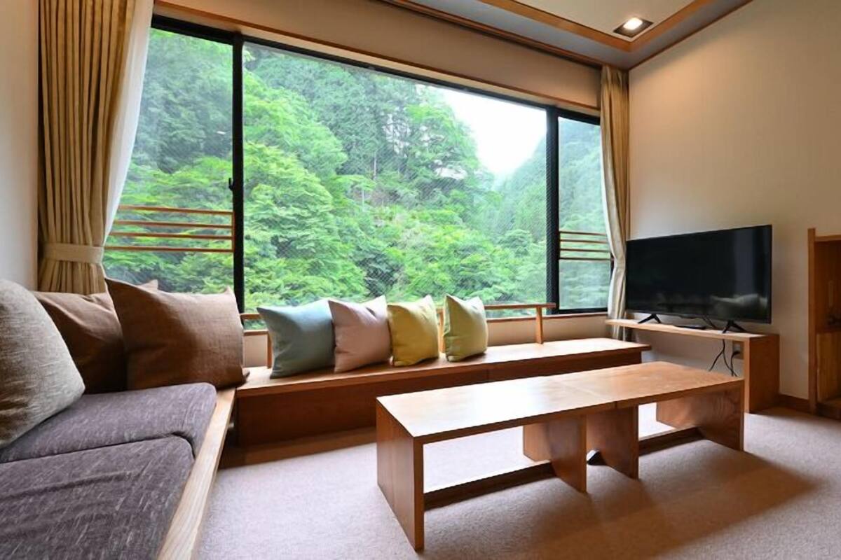 A living room with a sofa, a wooden coffee table, a TV on a stand, and a large window with a view of greenery.